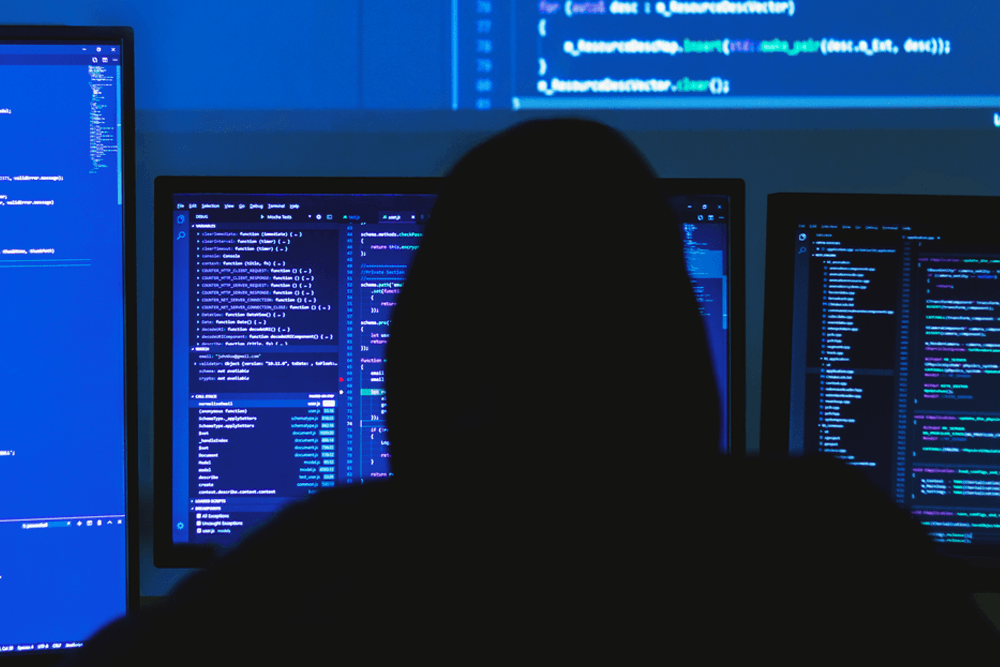 hacker in black hooded sweatshirt sits in front of multiple screens of code